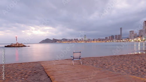 Playa de poniente desierta al atardecer en Benidorm, con el fato y las luces de los edificios en un día nublado con una silla abandonada en el camino de madera en la orilla photo