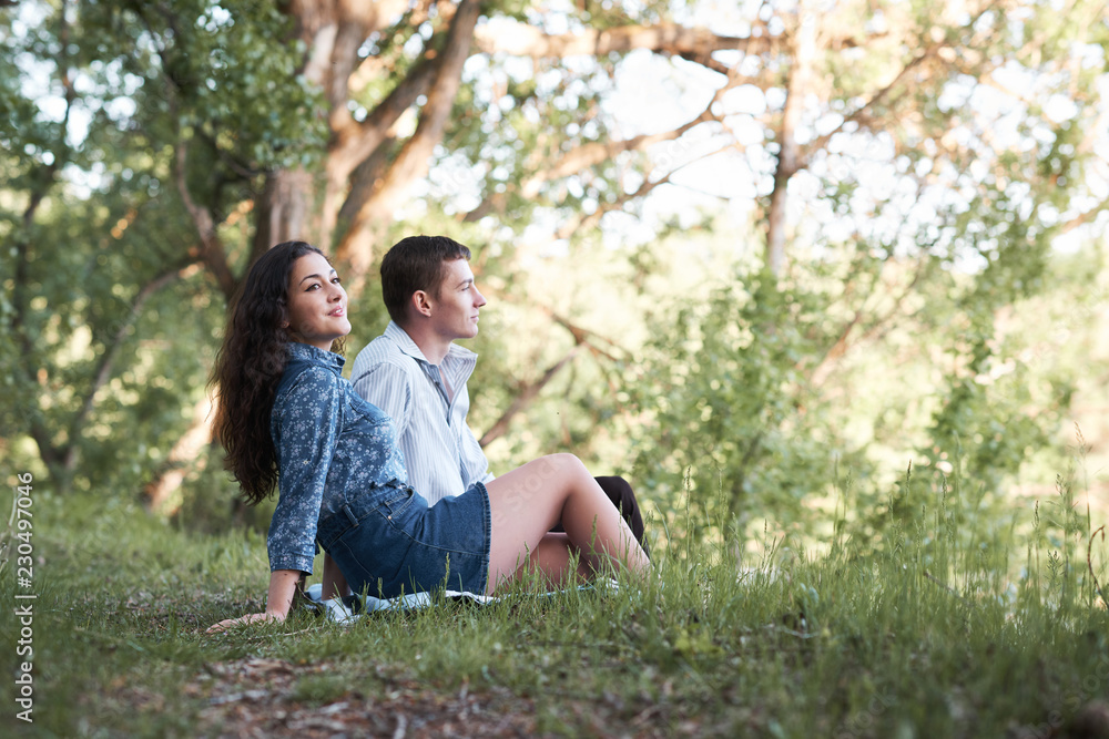 young couple sitting on the grass in the forest and looking on sunset, summer nature, bright sunlight, shadows and green leaves, romantic feelings