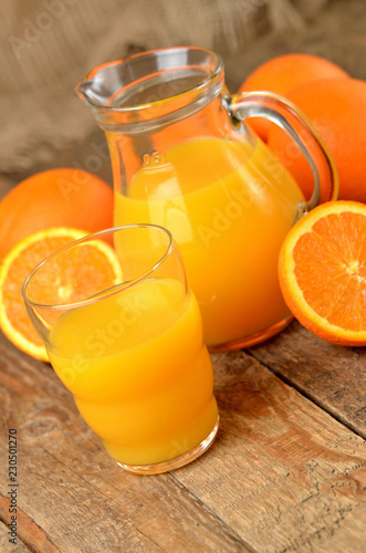 Orange juice in a glass, carafe with fresh juice and fresh oranges in the background on wooden table - vertical photo