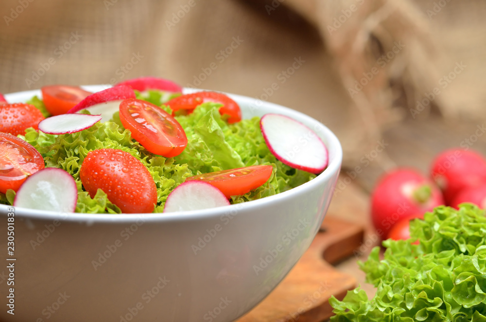 Close-up of green salad Lollo Biondo with tomatoes and radishes in a white bowl on wooden table, a pitcher of oil in the background - vertical photo Close-up of green salad Lollo Biondo with tomatoes and radishes in a white bowl on wooden table, a pitcher of oil in the background - vertical photo