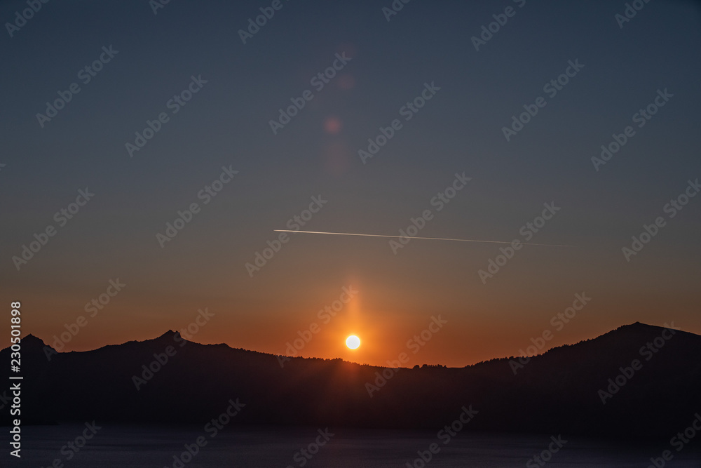 Scenic sunset over Crater Lake, Oregon. Sun setting down with a jet plane trail over it