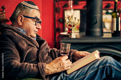 Jewish senior with glasses sitting in the armchair reading a torah book and drinking kosher wine