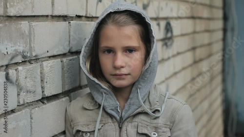 Sad young girl looking at camera with pain and reproach against grey brick wall. Difficult child wearing hooded jacket close up portrait outdoors.