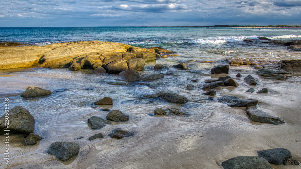 Large rocks and boulders in calm shallow sea water soft waves blue sky with clouds, wide image