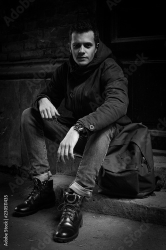 A young man with a hipster hanging on an outer garment on the stairs. Hoodshavy man in a wool jacket and high quality slippers with a backpack. Conceptual black and white photo.