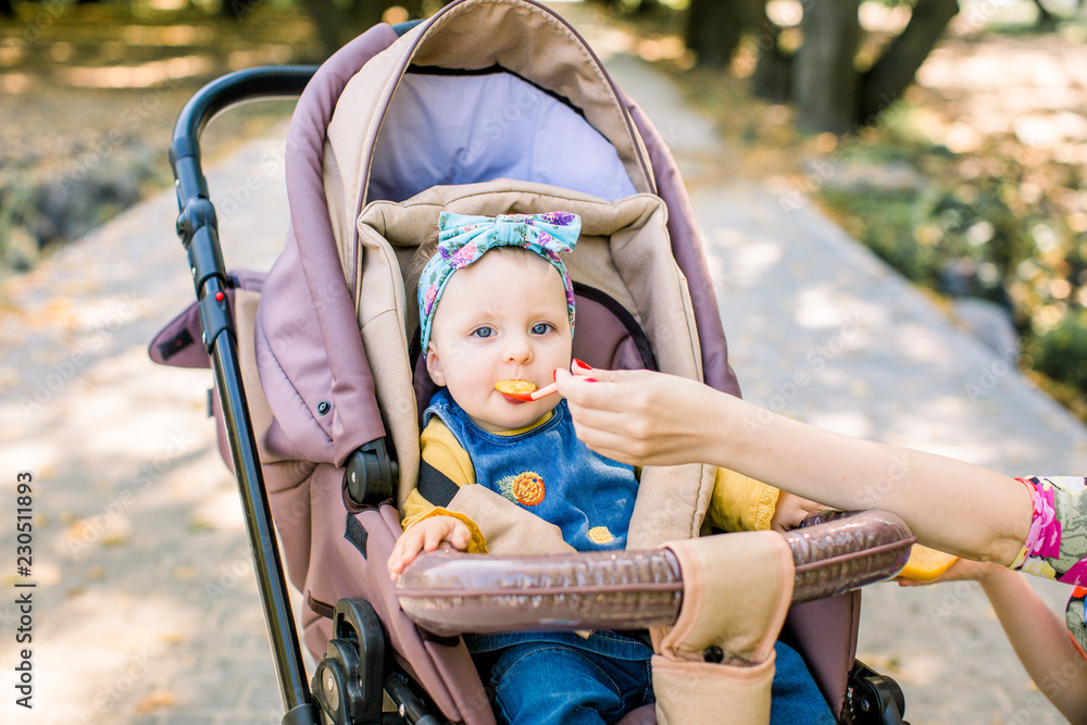 Young attractive mother feeding her cute baby girl, giving her first solid food, healthy vegetable pure from carrot with a plastic spoon sitting on baby stroller carriage and posing smiling Young attractive mother feeding her cute baby girl, giving her first solid food, healthy vegetable pure from carrot with a plastic spoon sitting on baby stroller carriage and posing smiling