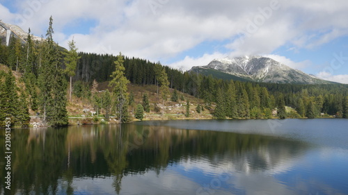 Strbske pleso and Tatra peaks visible from the back. A village located in the valley, from which tourists are moving to the Tatras. Colorful waters of a mountain pond, blue sky and unending peaks. © centryfuga