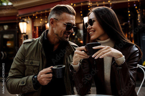Cafe. Couple. Love. Autumn. Man and woman in warm casual clothes and sun glasses are drinking coffee, talking and smiling while sitting in the cafe outdoors