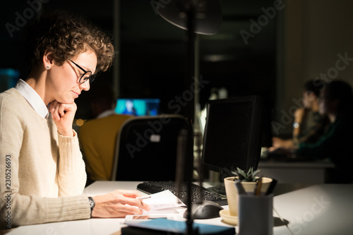 Side view portrait of young woman working late in dark office reading documents at table  copy space