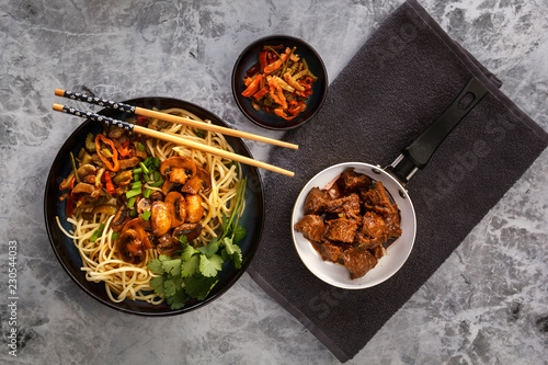 Chinese noodles with mushrooms, fried meat in a pan, vegetables and herbs are on the gray table. Top view