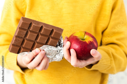 A child in a yellow sweater holding an apple and chocolate. Choosing between a healthy and unhealthy food