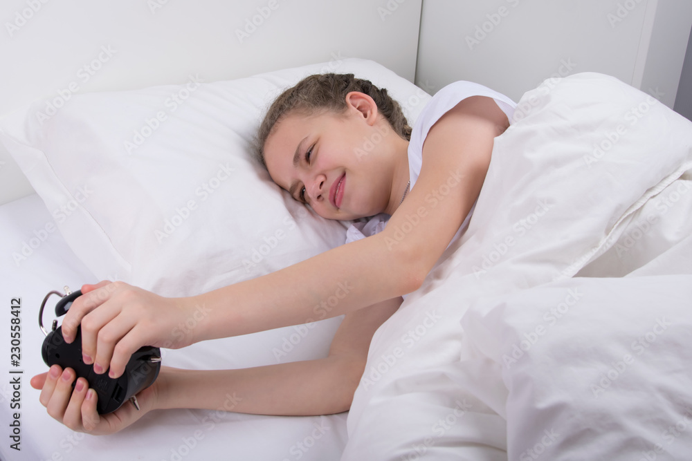 the girl, in a white bed, woke up in a good mood and looks at the alarm clock the girl, in a white bed, woke up in a good mood and looks at the alarm clock