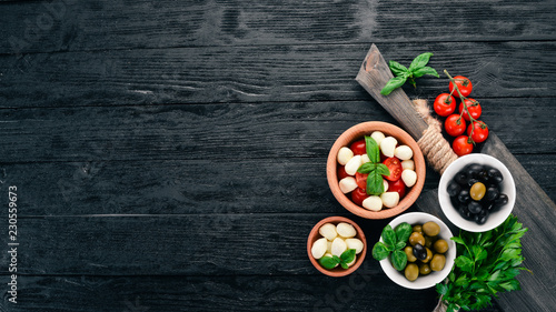 Ingredients for Italian caprese salad. Mozzarella cheese, cherry tomatoes, basil leaves, olives, oil, pepper. On a black wooden background. Free space for text.