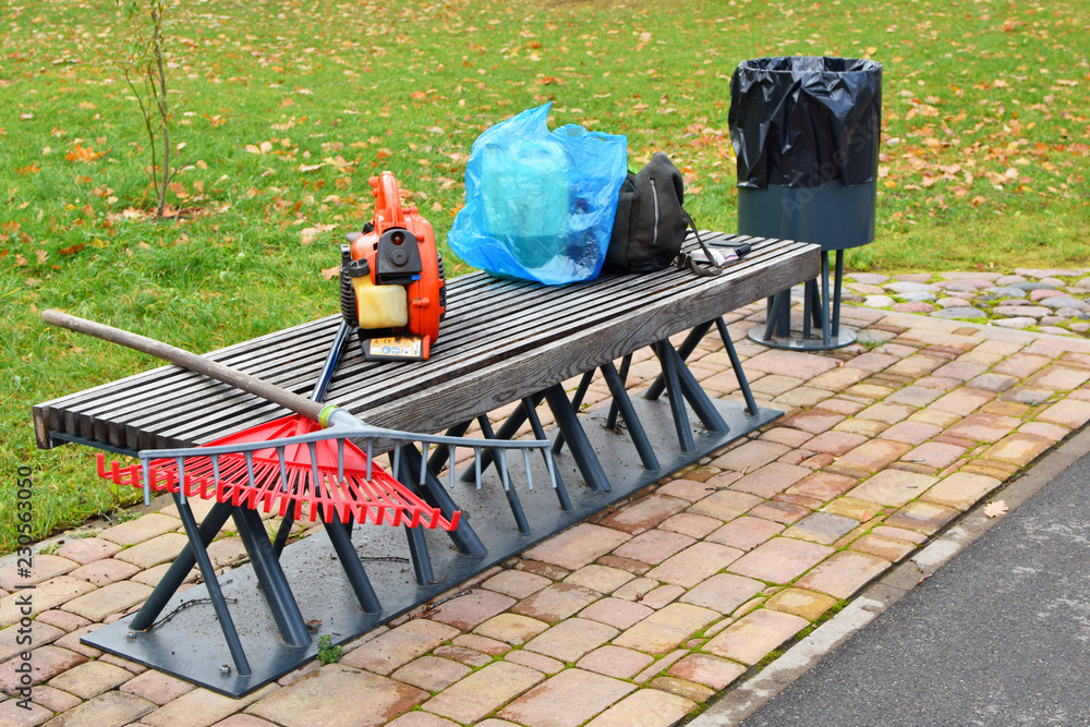 Φωτογραφία Worker equipment for cleaning fallen autumn leaves - rakes, leaf blower, fuel can on decorative wooden bench next to garbage bin outdoors in public park in autumn morning