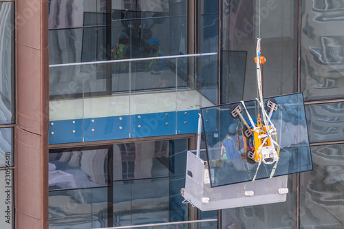 Construction workers wearing helmets on a suspended platform lifting a glass window frame.Glass installation or replacement on a high rise building