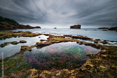 Vue sur un rocher en pose longue depuis la plage avec en premier plan un bassin translucide photo