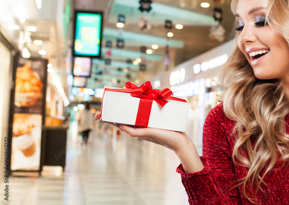 Laughing woman in Santa hat with Christmas gift box on shopping mall background. Christmas and New Year sale and offer concept Laughing woman in Santa hat with Christmas gift box on shopping mall background. Christmas and New Year sale and offer concept
