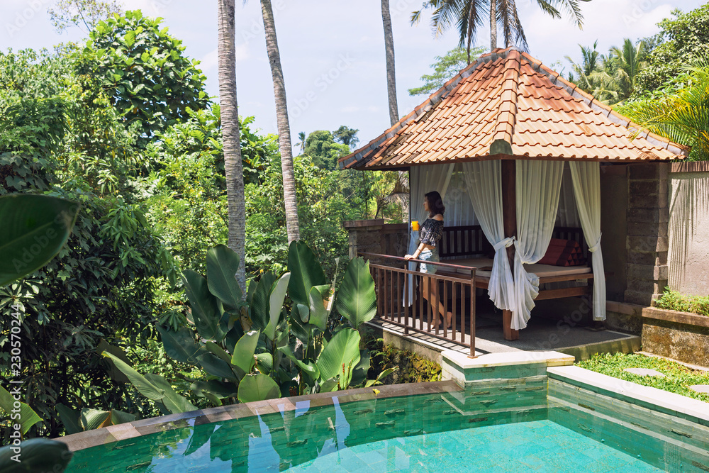 Φωτογραφία A young woman with a glass of juice in her hands is standing by the private pool against the backdrop of a romantic gazebo and tropical plants