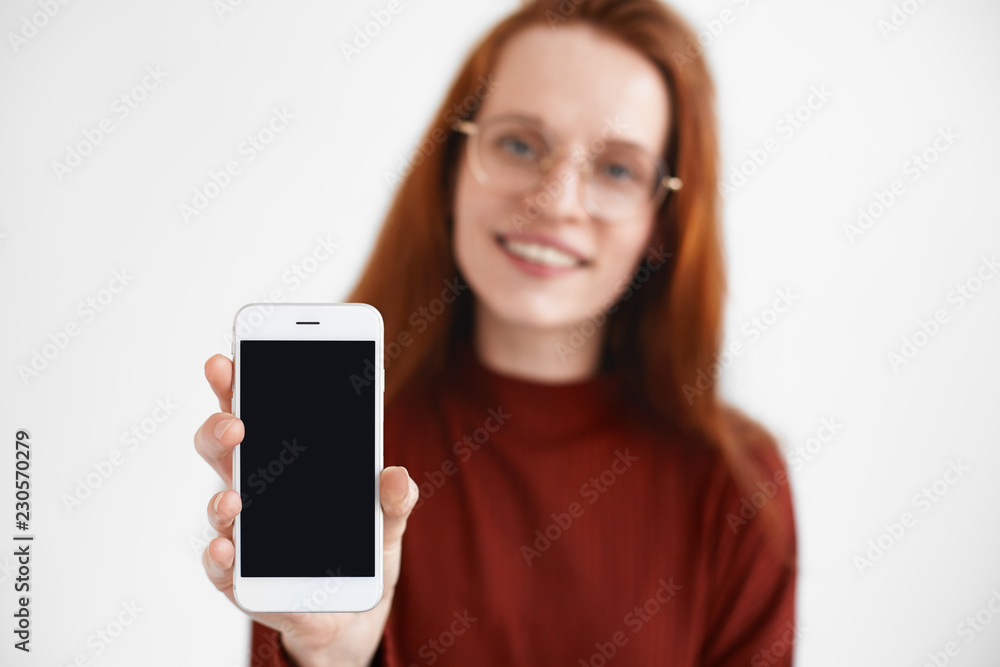 Cheerful young redhead female in glasses smiling, reaching out hand, holding generic mobile phone with black empty display with copy space for your text or advertising content. Selective focus