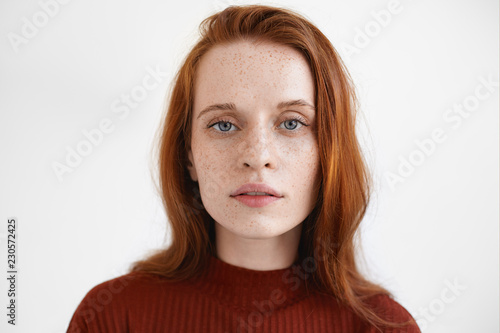 Gorgeous blue eyed red haired young European lady in maroon turtleneck looking at camera with calm facial expression, posing isolated against blank studio wall background with copyspace for your text photo