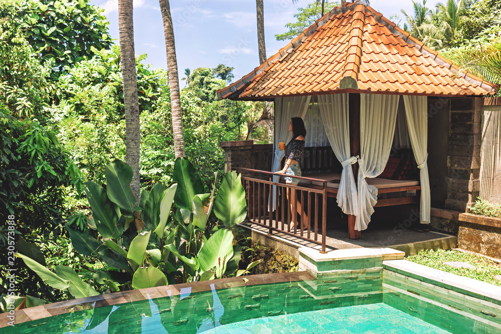 A young woman with a glass of juice in her hands is standing by the private pool against the backdrop of a romantic gazebo and tropical plants. A girl enjoys relaxing at a villa in Bali in Ubud.