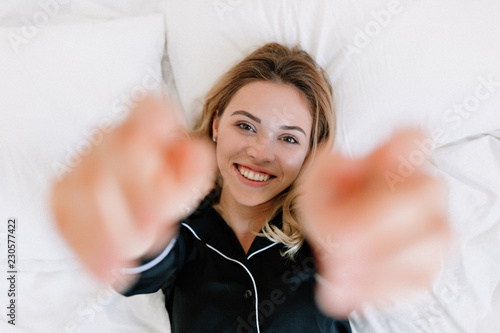 Portrait of wonderful blond girl on bed in modern apartment in the morning. She pulls hands in frame  withl ovely smile and looks satisfied photo