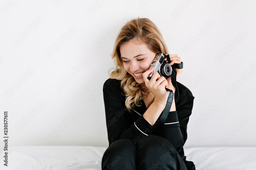Enchanting blonde woman in black night suit has fun with retro camera on light background with smile. Cute european girl in pajamas enjoying good morning and laughing. Enchanting blonde woman in black night suit has fun with retro camera on light background with smile. Cute european girl in pajamas enjoying good morning and laughing.