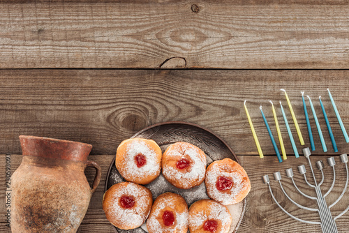 top view of sweet donuts, clay jug and menorah with candles on wooden tabletop, hannukah celebration concept photo