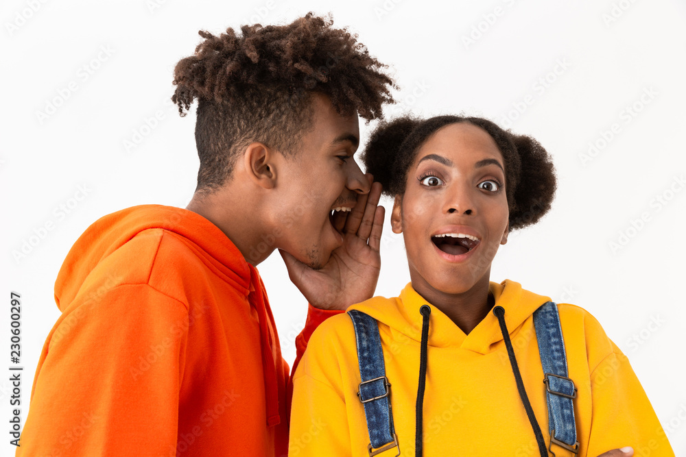 Fototapeta premium Photo of joyful african american man talking secrets in ear of his sister, isolated over white background