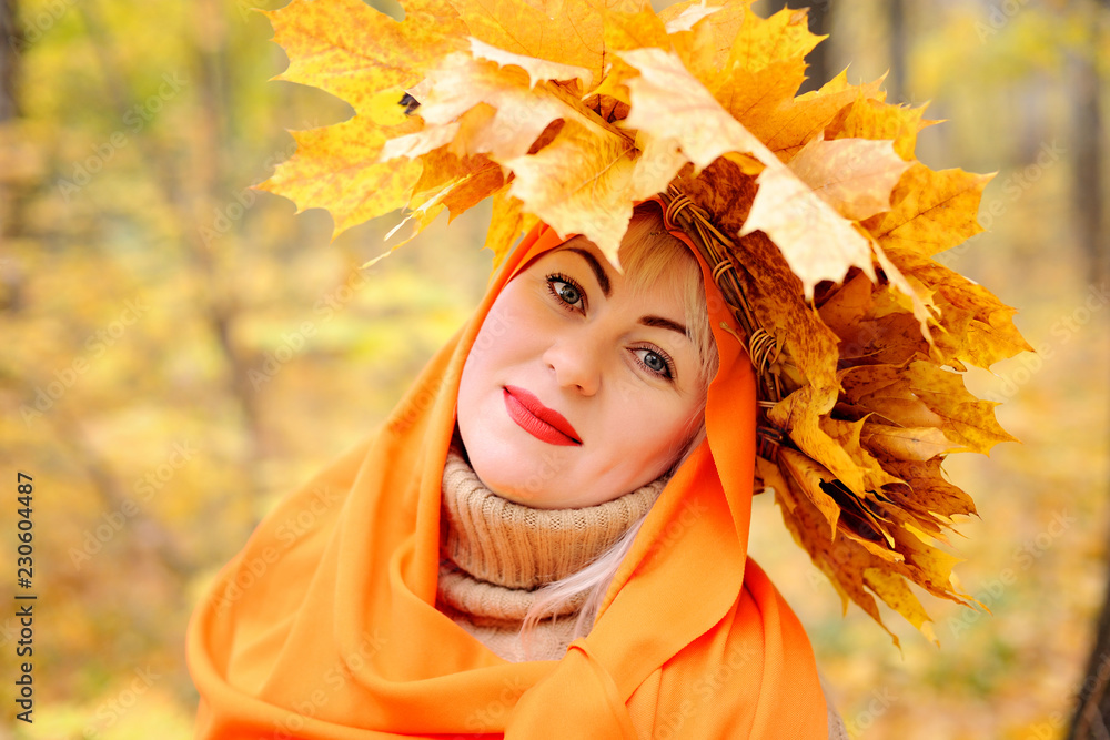 A large portrait of a beautiful middle-aged blonde in a wreath of autumn leaves, which stands in an orange scarf in the forest on a yellow background, looks at the camera and smiles. Plus size woman A large portrait of a beautiful middle-aged blonde in a wreath of autumn leaves, which stands in an orange scarf in the forest on a yellow background, looks at the camera and smiles. Plus size woman