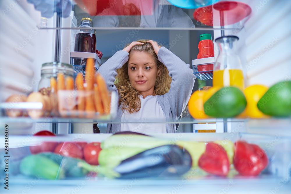 Confused woman choosing what to eat. Hands on head. Picture taken from the inside of fridge full of groceries. Confused woman choosing what to eat. Hands on head. Picture taken from the inside of fridge full of groceries.