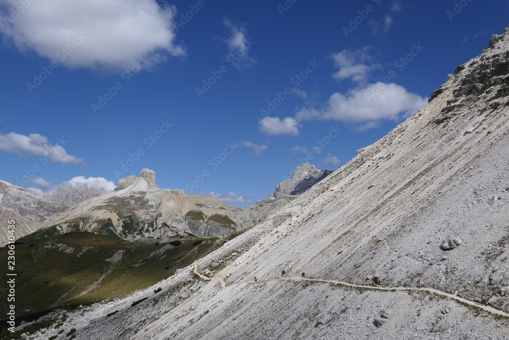 Alpine landscape. A narrow trail leads along a slope. It is a mountainous landscape in summer. The steep slope has a 45 degree angle and is very even. Blue sky and white clouds at lunchtime.