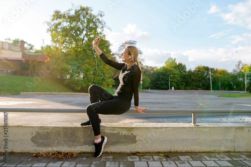 Side view of beautiful blonde woman in sportswear making selfiies while sitting on the handrail during morning training in the park.
