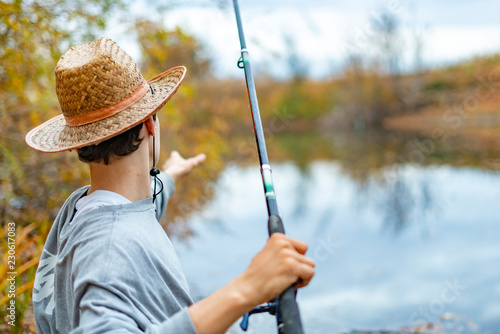 young man in hat sitting near lake and install set up and adjust fishing rod f