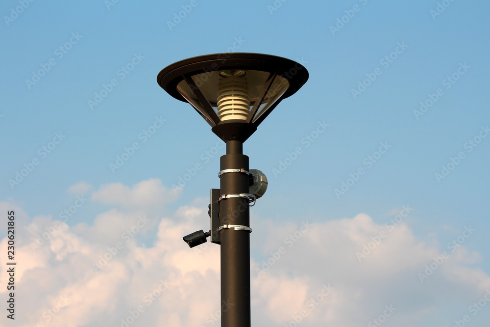 Modern metal black street lamp post with LED lightning and small surveillance camera with motion detector mounted on one side on cloudy blue sky background Modern metal black street lamp post with LED lightning and small surveillance camera with motion detector mounted on one side on cloudy blue sky background