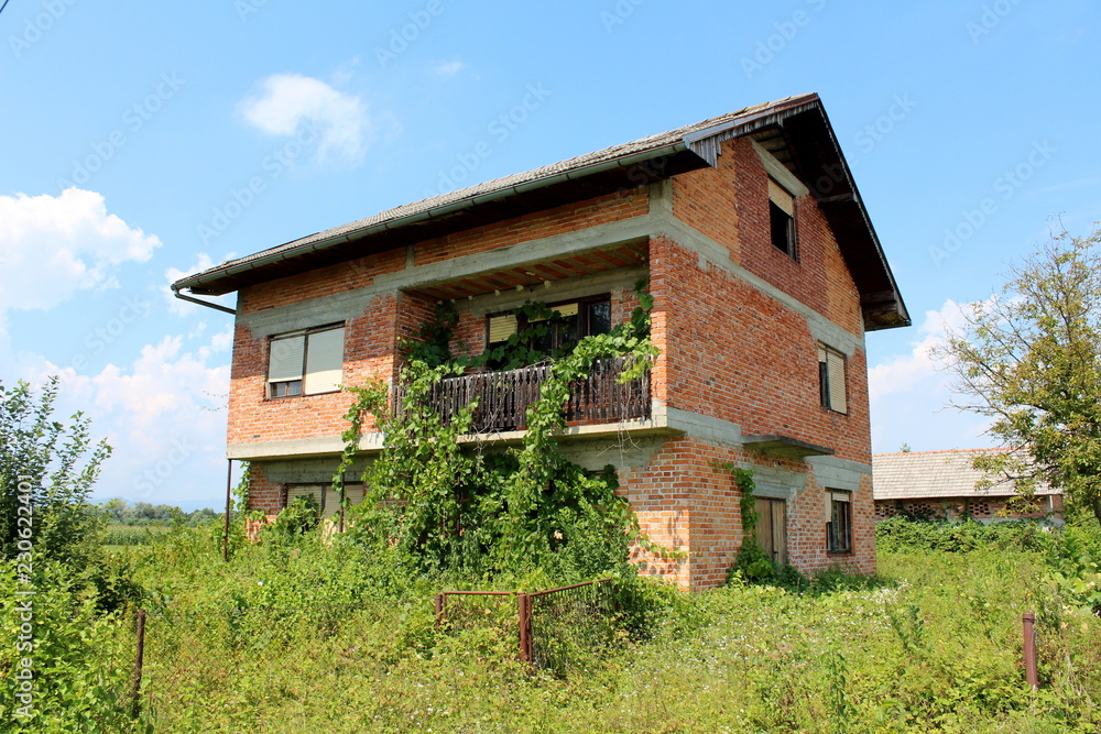Red bricks abandoned family house with broken windows and front wire fence overgrown with high uncut grass, trees and crawler plants with backyard building and cloudy blue sky in background on warm su Red bricks abandoned family house with broken windows and front wire fence overgrown with high uncut grass, trees and crawler plants with backyard building and cloudy blue sky in background on warm su