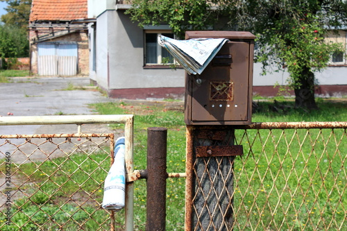 Rusted broken mailbox with faded newspaper mounted on wooden pole behind partially rusted wire and iron fence with abandoned family house and dilapidated garage in background