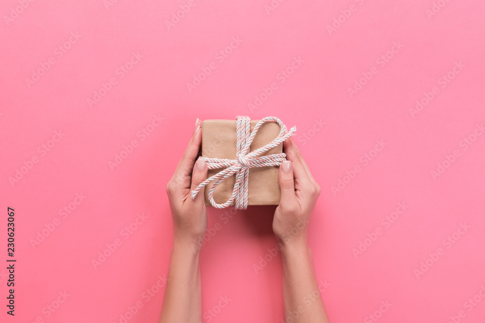 Woman hands give wrapped valentine or other holiday handmade present in paper with pink ribbon. Present box, decoration of gift on pink table, top view with copy space Woman hands give wrapped valentine or other holiday handmade present in paper with pink ribbon. Present box, decoration of gift on pink table, top view with copy space