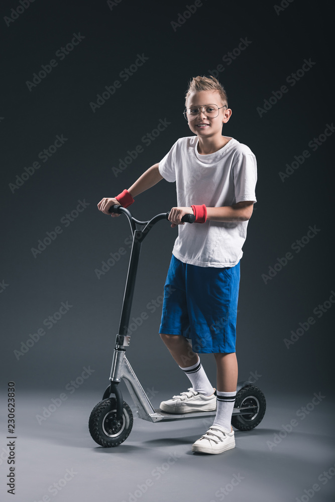 smiling stylish boy in eyeglasses on scooter looking at camera on grey backdrop smiling stylish boy in eyeglasses on scooter looking at camera on grey backdrop