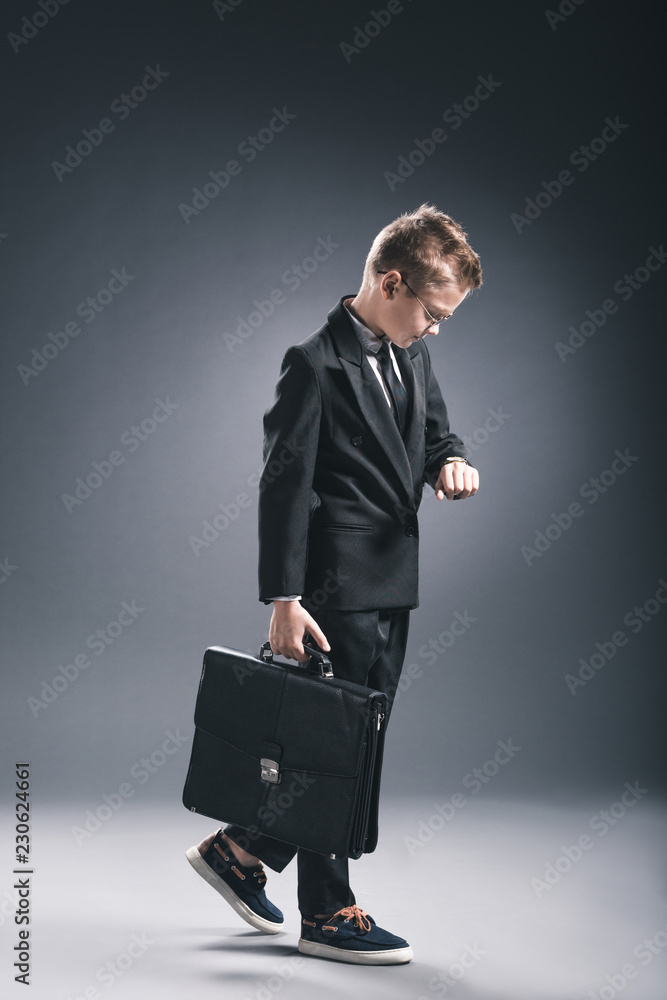 preteen boy in businessman suit and eyeglasses with suitcase checking time on dark background preteen boy in businessman suit and eyeglasses with suitcase checking time on dark background