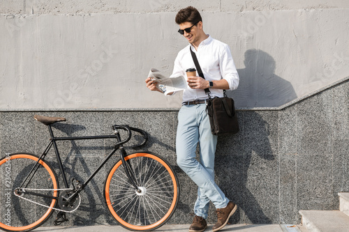 Photo of young man 30s wearing sunglasses, drinking takeaway coffee and reading newspaper, while standing with bicycle along wall outdoor