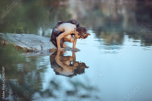 Caveman boy sitting on the rock and looking at him self in the water reflection in lake. Evolution survival concept. Creative art fantasy photo photo
