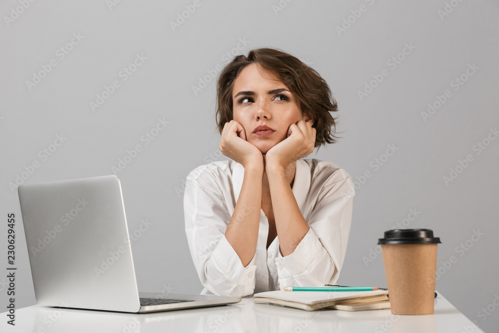 Thinking young woman sitting at the table isolated over grey background using laptop computer. Thinking young woman sitting at the table isolated over grey background using laptop computer.