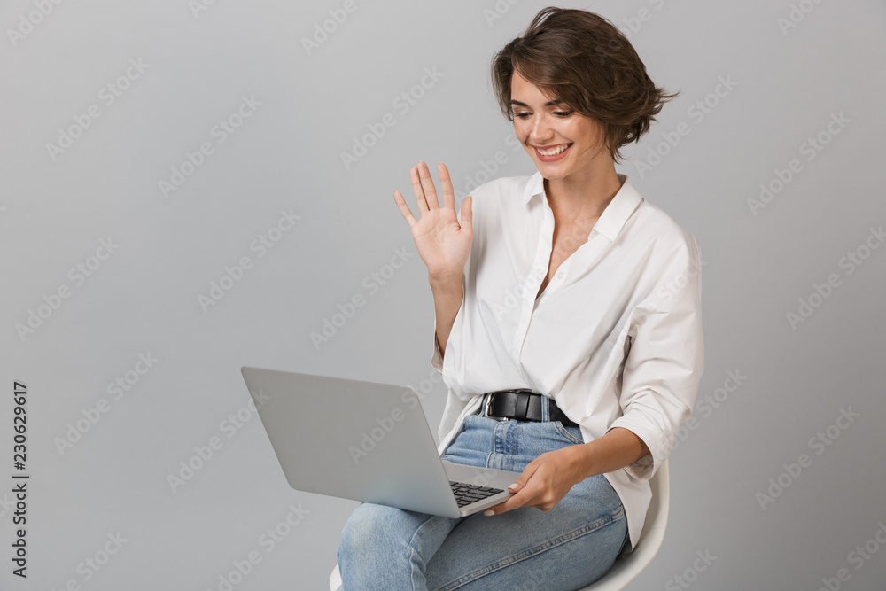 Happy young woman sitting on stool isolated over grey background holding laptop computer waving. Happy young woman sitting on stool isolated over grey background holding laptop computer waving.
