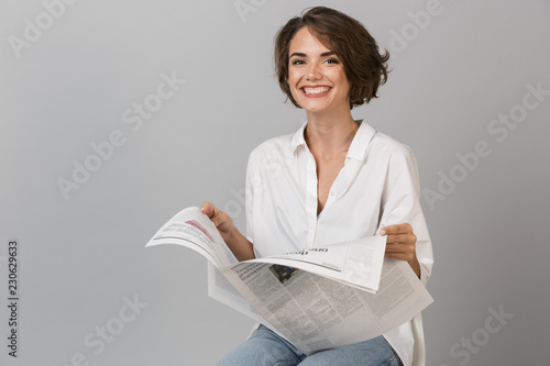 Happy young woman sitting on stool isolated over grey background holding newspaper reading. photo