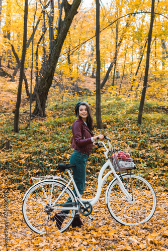 happy fashionable woman in stylish leather jacket and beret carrying bicycle in forest