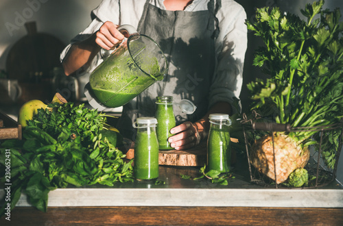 Making green detox take-away smoothie. Woman in linen apron pouring green smoothie drink from blender to bottle surrounded with vegetables and greens. Healthy, clean eating, weight loss food concept