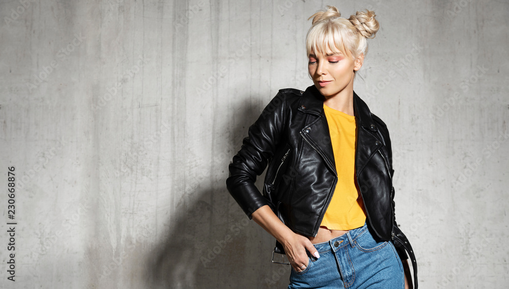 Naklejka premium Stylish woman wearing leather jacket. Young blond-haired female posing in studio on cement background. Modern fashion concept.