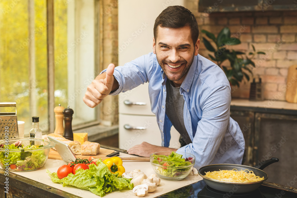 Portrait of young smiling man preparing delicious and healthy food in the home kitchen on a sunny day. Using tablet computer for searching recipes. Thumbs up. Portrait of young smiling man preparing delicious and healthy food in the home kitchen on a sunny day. Using tablet computer for searching recipes. Thumbs up.