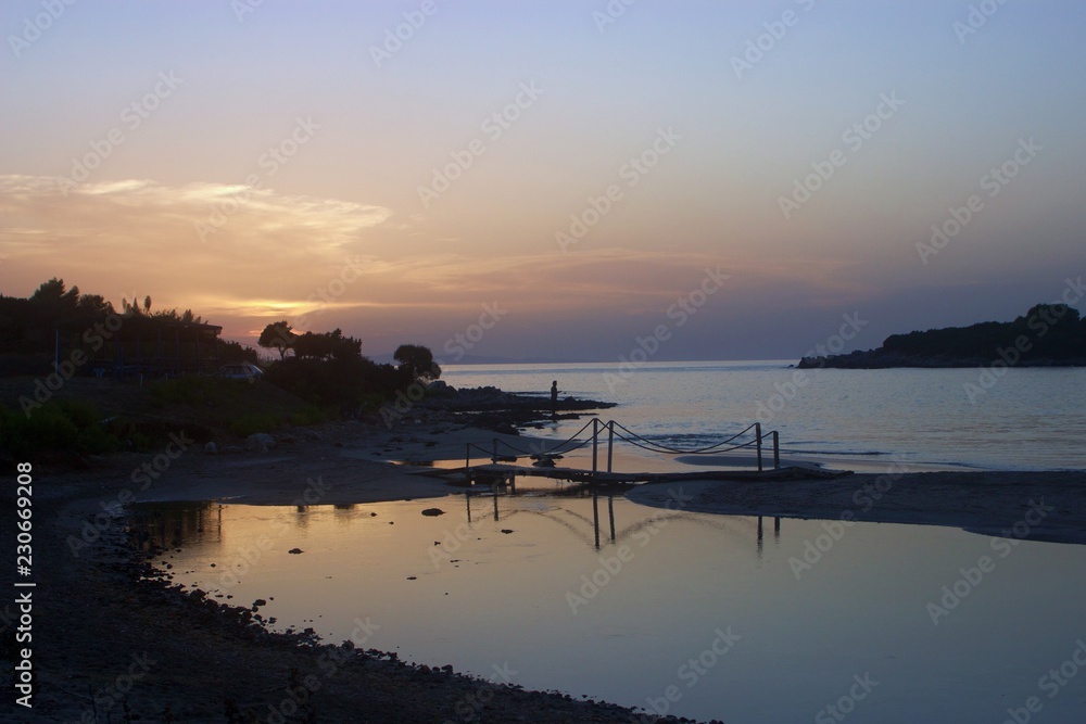 Fototapeta premium Ormos odyssea beach - kerenza near Parga at sunset, with a creek and a small bridge, silhouette of a fisherman fishing in the background